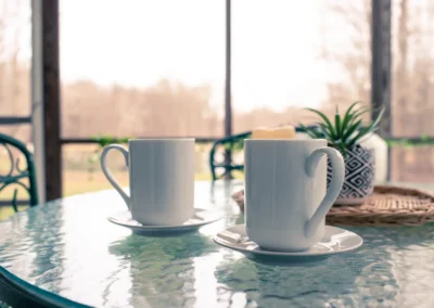 Two white mugs on a glass patio table overlooking a wooded area.