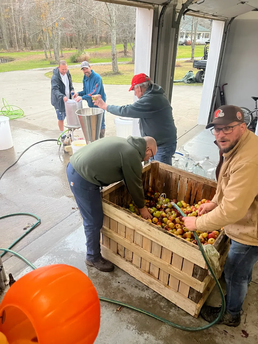 A group of people working together in a garage to process apples, with one person sorting apples in a wooden crate, while others are using equipment and preparing for a task.