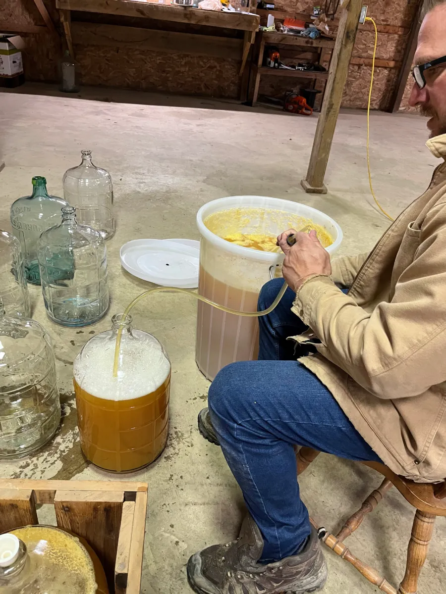 A person sitting on a chair in a barn, working with fermentation equipment and glass containers filled with liquid.