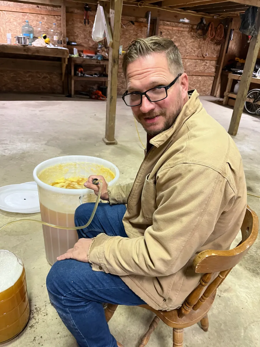 A man sitting on a wooden chair in a workshop, wearing a beige jacket and glasses, while working with a large container of liquid.